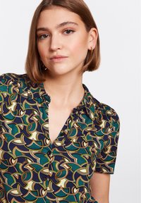 Kurze Ärmelbluse mit einem abstrakten Muster in Grün, Marineblau und Beige, mit Knopfverschluss und rundem Kragen, aus leichtem Material gefertigt.