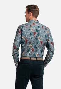 Langsleeve shirt in blauw met een bloemenpatroon met teal, rode en beige bloemen. Draagt met donkere broek en een geweven riem.