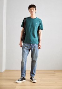 Teal short-sleeve t-shirt, relaxed fit, worn with light blue jeans and black sneakers. Brown shoulder bag slung across the body.