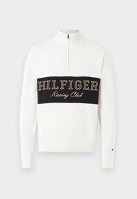 Balts džemperis ar ķēdītes apkakli, ar ribotām aprocēm, kas rotāts ar melnu horizontālu joslu, uz kuras izšūts "HILFIGER Racing Club" tan krāsā. Mīksta adīta tekstūra.
