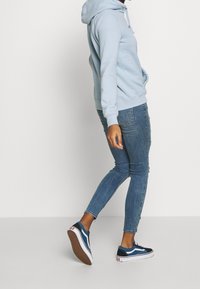 Felpa con cappuccio azzurro chiaro con tasca a marsupio, abbinata a jeans blu aderenti e sneakers blu navy con suola bianca. Vista laterale dell'outfit.