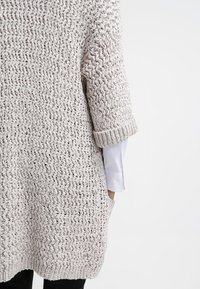 Ljust beige stickad tröja med lös passform, texturerat mönster, rullade ärmar och sidofickor. Mjukt material och avslappnad design.