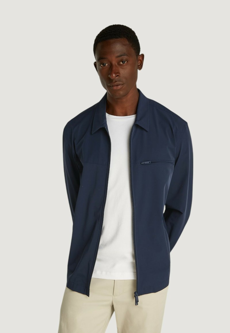 Giacca leggera di colore blu navy con zip davanti, colletto a punta e taschino sul petto. Indossata sopra una t-shirt bianca e pantaloni beige.