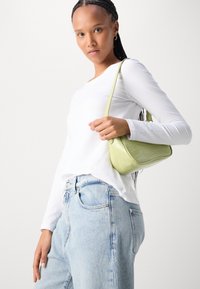 Green crocodile-patterned handbag with a curved base, small handle, and smooth texture, worn over a white long-sleeve top and light blue jeans.