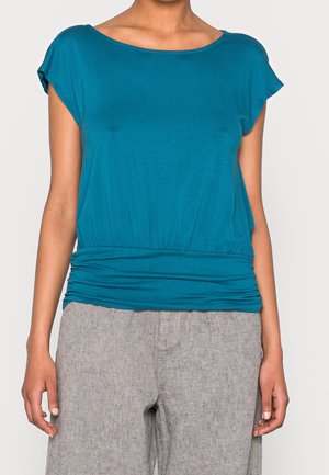 Woman wearing a teal short-sleeve top with a gathered waistband paired with loose-fitting light gray pants.