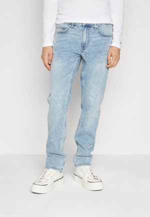 Personne portant un jean droit bleu clair, une chemise blanche à manches longues et des baskets blanches, debout devant un fond uni.