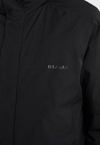 Sort isoleret jakke med en glat tekstur. Har en høj krave og et præget logo "BERLUTI BAZAAR" på forsiden.