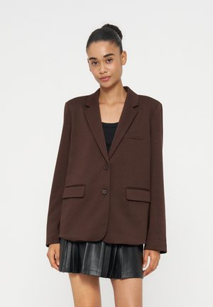 Femme aux cheveux en chignon portant un blazer marron foncé oversize sur un haut noir et une jupe en cuir plissée noire, debout devant un fond blanc.