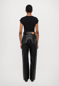 Pantaloni din piele neagră cu picioare largi, cusături albe și buzunare aplicate, asortați cu un tricou negru mulat, văzuți din spate.