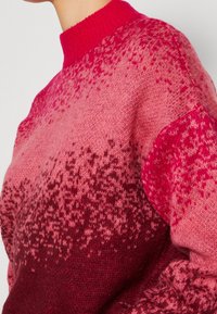 Gros plan d'un pull tricoté rouge et rose avec un motif moucheté en dégradé et un col montant côtelé.