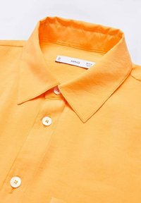 Camicia collare arancio brillante con bottoni bianchi e un'etichetta del marchio visibile all'interno del colletto, stesa su una superficie bianca.