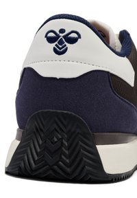 Sneakerhæl i sort gummi med zigzag-tryk, mørkeblå ruskind og mesh, hvide læderdetaljer, med et broderet logo nær toppen.
