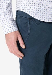 Blue textured pants with traditional pockets, paired with a white shirt featuring a blue pattern. The hand rests on the pant pocket.