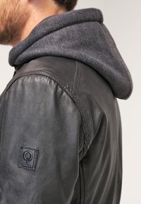 Giubbotto di pelle nera con cappuccio grigio, caratterizzato da una superficie a costine, cuciture in evidenza e una patch con logo sulla manica.
