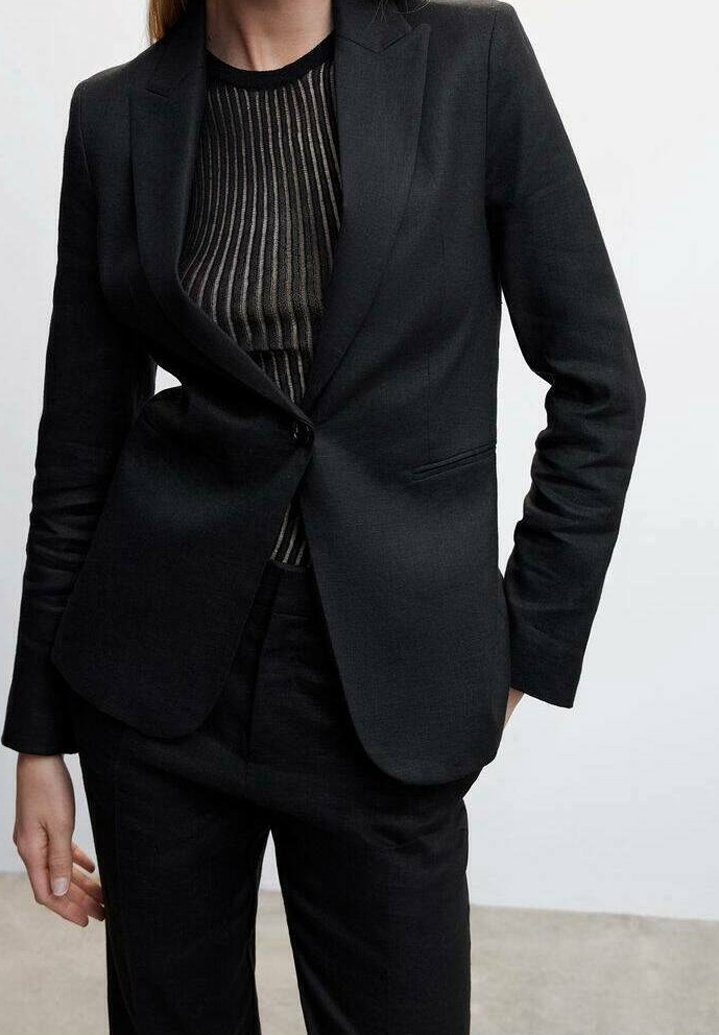 Personne portant un blazer noir ajusté sur un haut rayé noir et gris, associé à un pantalon noir, debout devant un fond uni.