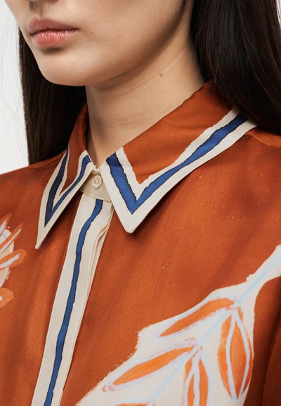 Γυναίκα που φοράει πουκάμισο χρώματος σκουριάς με λευκό και μπλε ριγέ γιακά και αφηρημένα πορτοκαλί και λευκά λουλουδένια σχέδια.