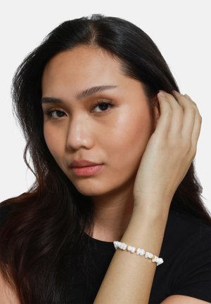 Weißes Perlenarmband am Handgelenk, mit unregelmäßigen Formen, einer glatten Textur und kleinen goldenen Akzenten. Modell trägt ein schwarzes Oberteil.