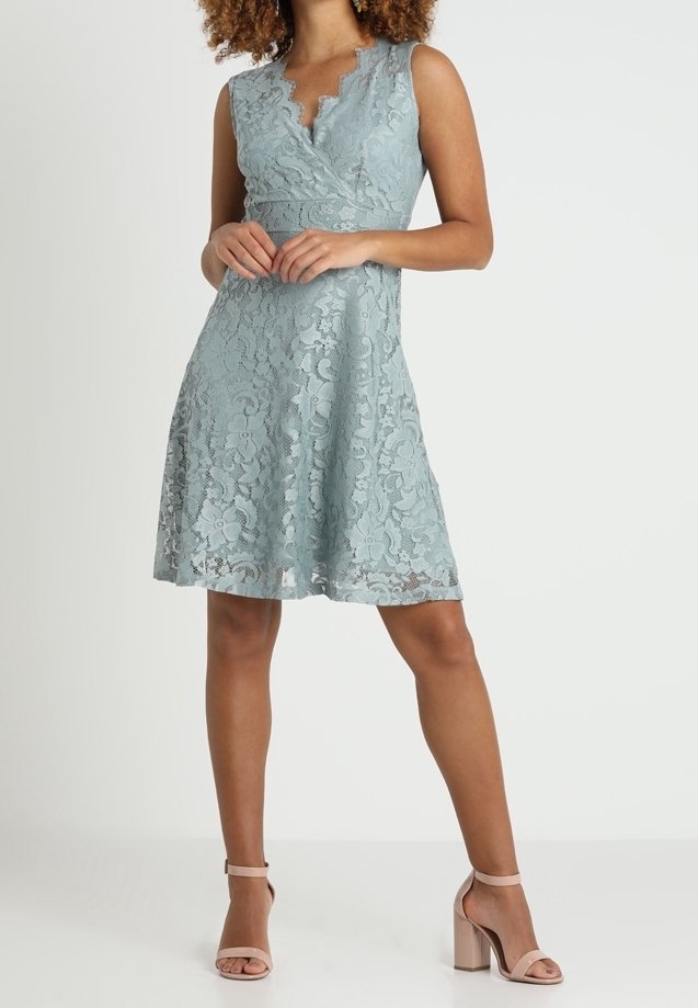 Femme portant une robe sans manches en dentelle bleu clair avec un col en V festonné, associée à des talons blocs nude à bride à la cheville, debout, mains jointes.