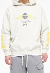 Krémová mikina s kapucí, přední kapsou a stahovací šňůrkou, s grafickým potiskem křídel a textu v černé a žluté barvě na přední straně a rukávech.