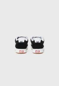 Sorte semskede lavtoppede sneakers med hvite detaljer, som har et rutet mønster på hælen og en rød logoetikett på den hvite gummisål.