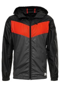 Veste de pluie noire avec un motif texturé, accent diagonal rouge sur la poitrine, capuche ajustable et fermeture éclair frontale complète.