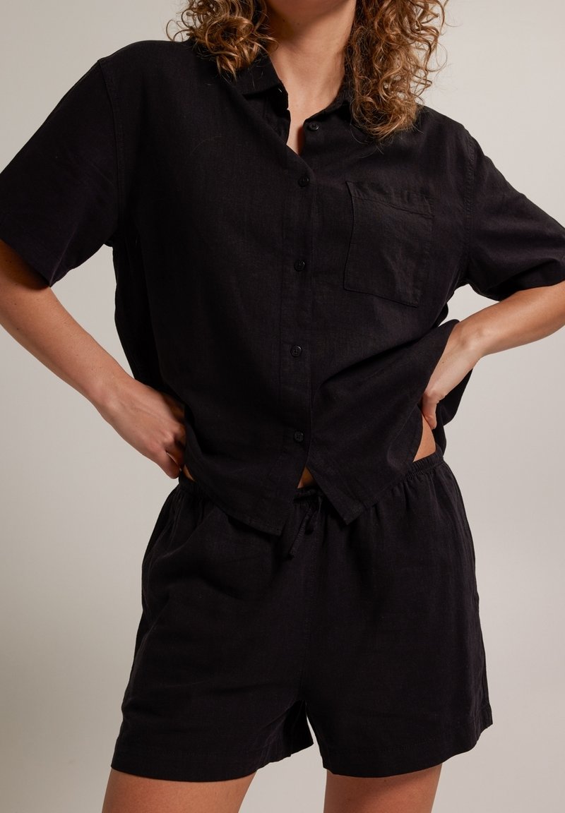 Černá lněná košile s krátkým rukávem, s knoflíkovým zapínáním vpředu a jednou kapsou na prsou, spárovaná s odpovídajícími černými šortkami. Pohodlný, volný střih.