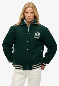 Groene wollen jas met een witte kraag, zilveren hardware en een logo aan de voorkant. Voorzien van gestreepte ribgebreide boorden en zoom, zijzakken voor opslag.
