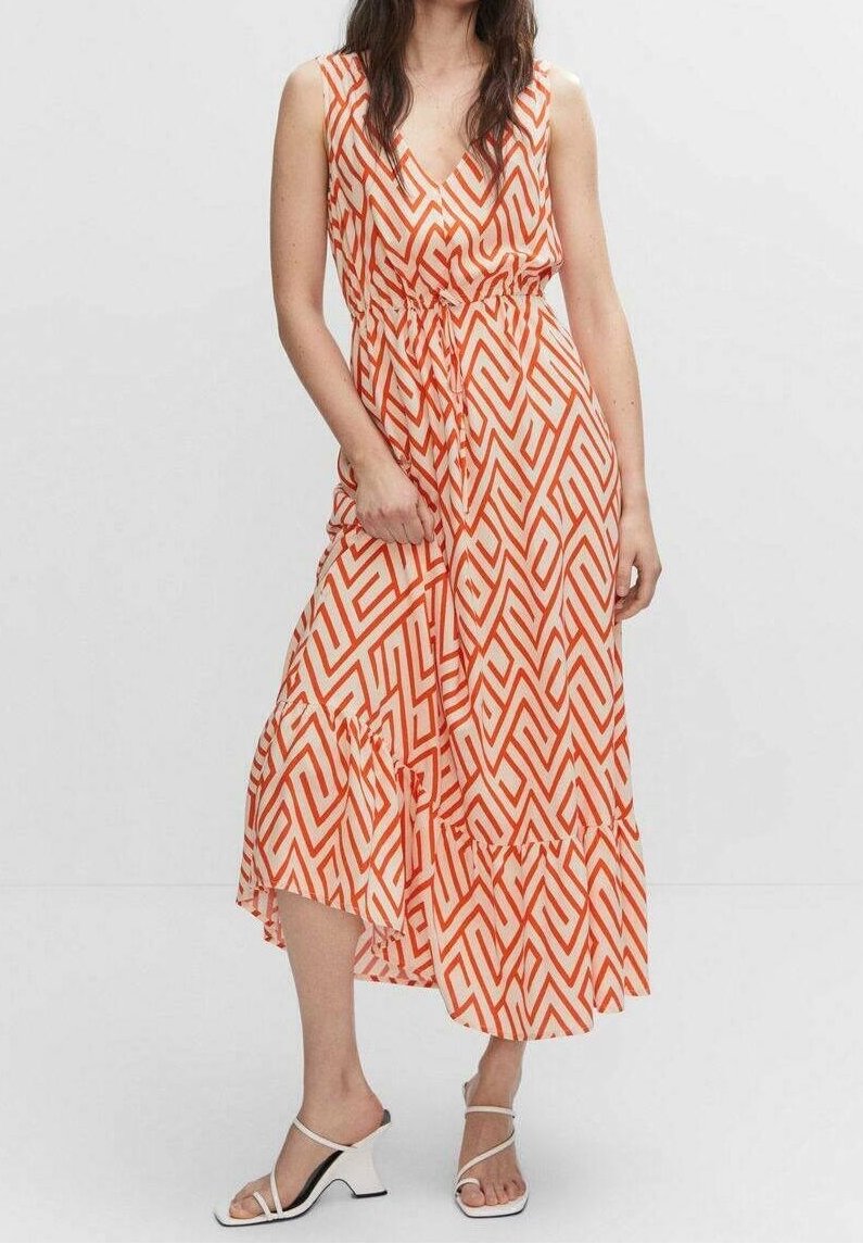 Femme portant une robe midi sans manches à imprimé géométrique orange et blanc avec des sandales blanches à talons, debout devant un fond uni.