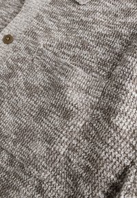 Gebreide cardigan in getextureerde lichtbruin en crèmekleurige tinten, met een klein borstvakje en een enkele bruine knoopsluiting.
