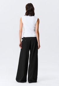 Haut blanc sans manches en tissu texturé, associé à un pantalon noir taille haute à jambes larges, avec poches arrière et détails à boutons.