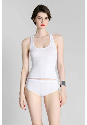 Femme aux cheveux courts et foncés portant un débardeur blanc et un slip, rouge à lèvres et vernis à ongles rouge, debout devant un fond gris uni.