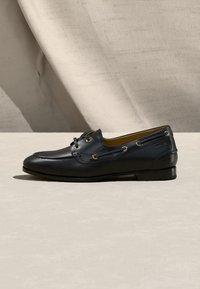 Mocassin pour homme en cuir noir avec détails en lacets et petit talon, présenté sur une surface beige avec un fond en tissu clair.