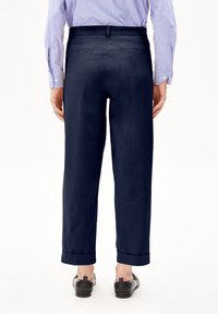 Pantaloni in cotone blu navy con una vestibilità comoda, dotati di un risvolto, tasche posteriori e indossati con scarpe nere. Maniche della camicia a righe visibili.