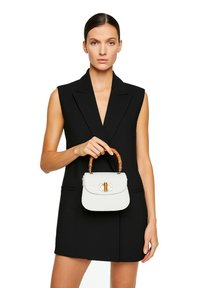 Borsa in pelle bianca con manico in bambù, dettagli in oro, forma rettangolare e chiusura a patta. Indossata da un modello in un vestito nero senza maniche.