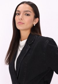 Fekete farmerblézer hegyes hajtókákkal, fehér inggel párosítva, ezüst láncnyakláncokkal. Simára fésült haj és egyszerű fülbevalók.