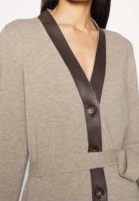 Béžový svetr s tmavě hnědým saténovým šálovým límcem, předními knoflíky a odpovídajícím páskem kolem pasu. Měkká textura s mírně přiléhavým střihem.