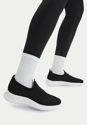 Feet wearing black slip-on sneakers with white soles and white ribbed socks, paired with black leggings against a plain background.