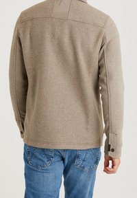 Brun texturerad jacka med en figursydd design, med en okdetalj på baksidan och knapppynt på ärmarna, kombinerad med blå jeans.