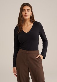 Schwarzes Langarmoberteil mit V-Ausschnitt, kombiniert mit braunen hochsitztenden Hosen. Der glatte Stoff und die taillierte Passform sorgen für einen eleganten, sauberen Look.