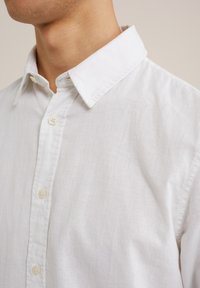 Witte linnen overhemd met een button-down kraag, voorzien van zichtbare knopen en een gestructureerde stof. Close-up weergave benadrukt de kraag en de stof.
