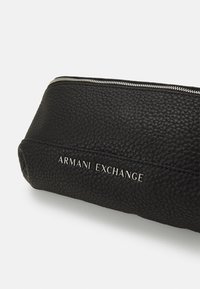 Zwart gestructureerd leren etui met zilveren rits, voorzien van de embossering van de merknaam "ARMANI EXCHANGE" aan de zijkant.