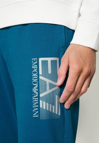 Närbild av en hand som vilar på blå byxor med vit vertikal rand och en vit sweatshirtärm synlig.