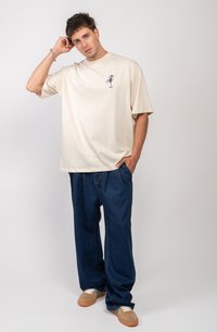 Camiseta oversized beige con un pequeño gráfico azul en el lado izquierdo del pecho, combinada con pantalones vaqueros oscuros y sueltos, y zapatillas marrones.