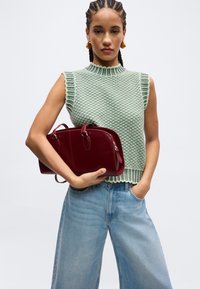 Γυναίκα με πλεξούδες που φοράει πράσινο αμάνικο πλεκτό τοπ και φαρδιά μπλε τζιν, κρατώντας με το ένα χέρι μια σκούρα κόκκινη τσάντα.