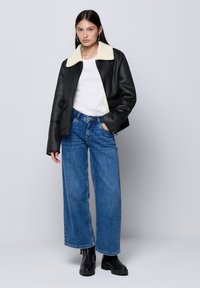 Giacca di pelle nera con colletto in pelliccia crema, indossata sopra una camicia bianca. Abbinata a jeans blu a gamba larga e stivaletti neri.