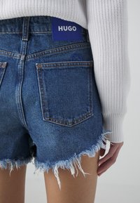 Short en denim bleu effiloché avec poches arrière, porté avec un pull blanc côtelé, affichant une étiquette bleue "HUGO" sur la ceinture.