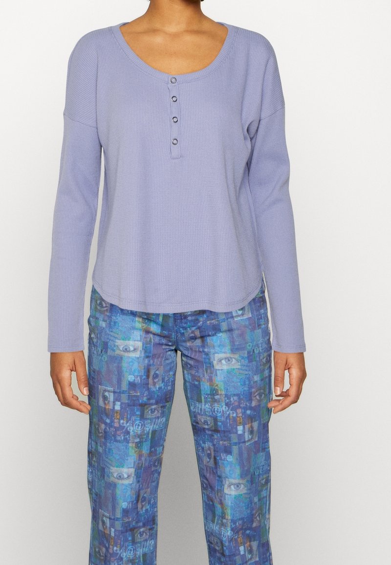Ljuslila långärmad skjorta med en rundad halsringning och tre knappar. Matchas med blå byxor med en kollage av ögon och abstrakta mönster.