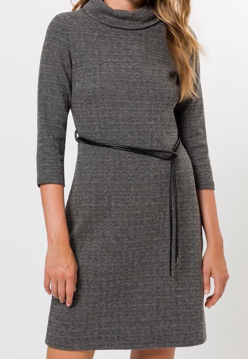 Robe en chevrons gris avec un col drapé, des manches trois-quarts et une ceinture noire soulignant la taille. Texture lisse, coupe ajustée.