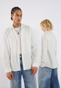 Chemise blanche texturée à ouverture frontale avec un col, des manches longues et des détails de boutons visibles, associée à un jean large bleu clair.