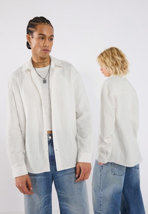 Witte, gemêleerde open voorkant blouse met een kraag, lange mouwen en zichtbare knoopdetails, gecombineerd met lichtblauwe wijdere jeans.